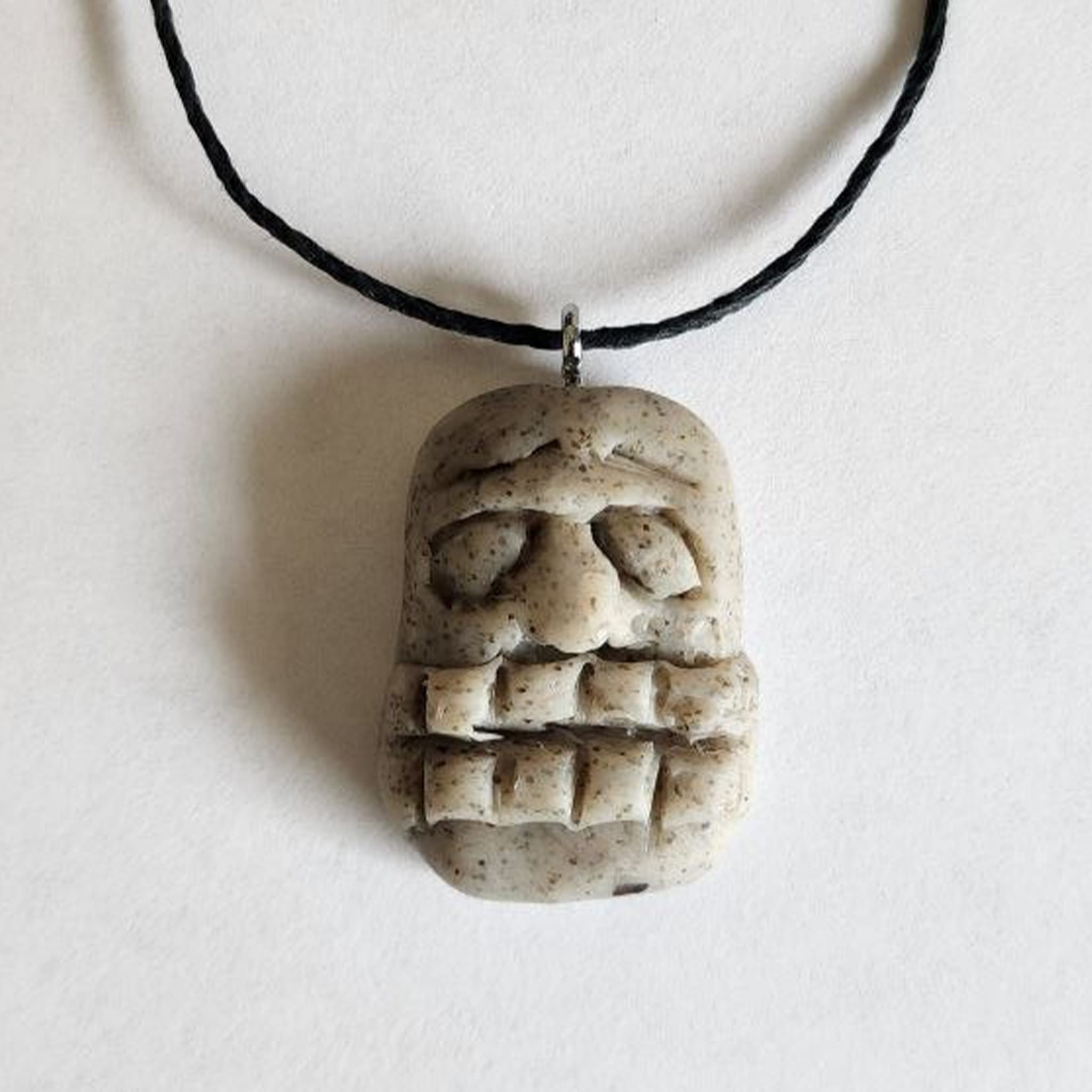 Handmade stone-textured ceramic pendant of a Taíno spirit face with a prominent nose, straight mouth, and horizontal line eyes on a black cord necklace, inspired by indigenous Puerto Rican petroglyphs.