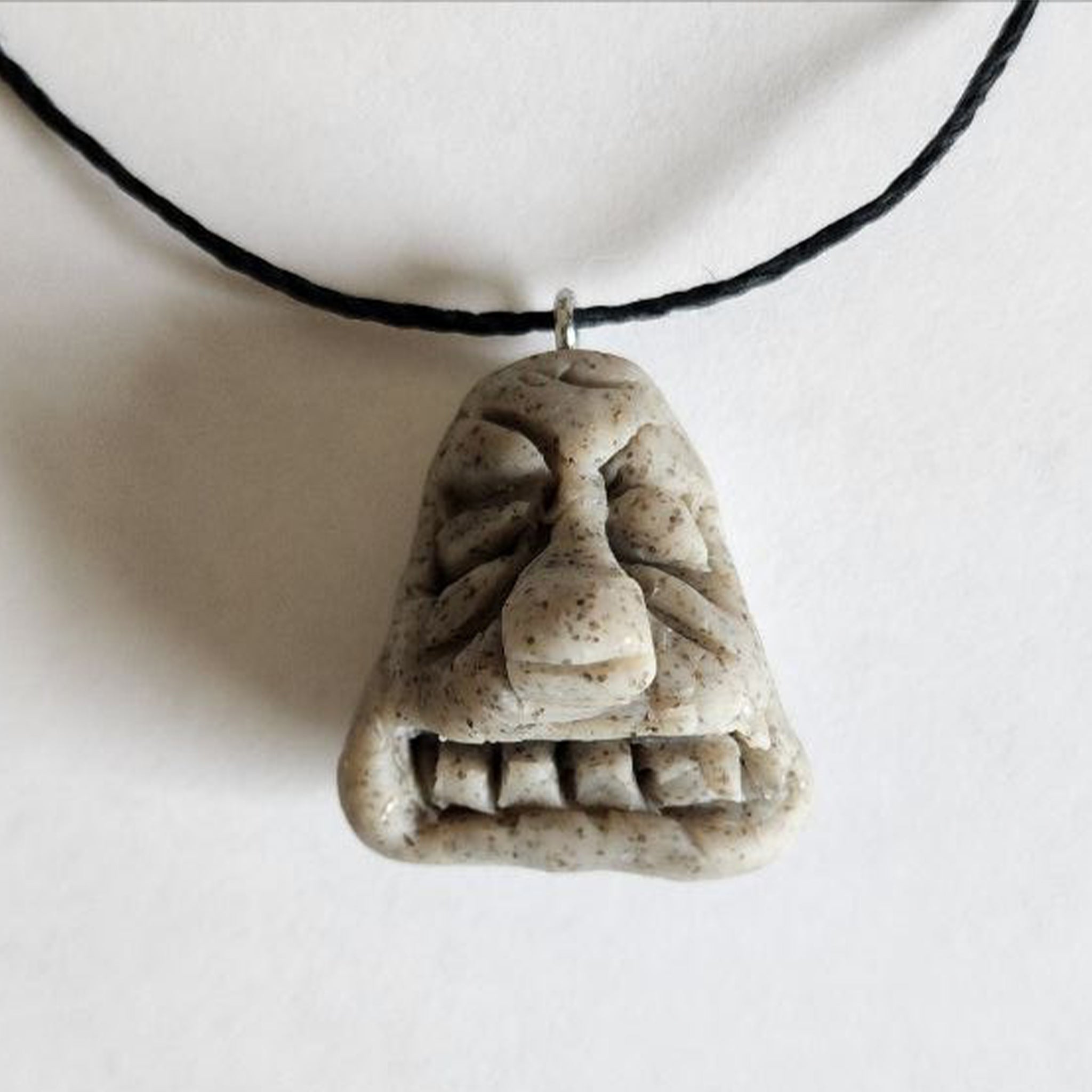 Handmade stone-textured ceramic pendant of a triangular Taíno spirit face with closed eyes, a big nose, and straight teeth on a black cord necklace, inspired by indigenous Puerto Rican rock art.