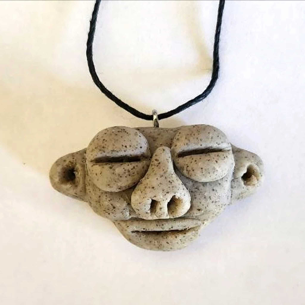 A triangular ceramic pendant featuring a Taíno Zemí spirit face with closed eyes and a prominent nose, handcrafted with a grey stone texture on a black adjustable cord.