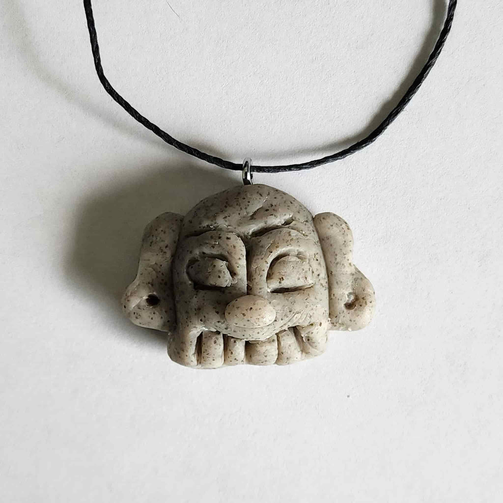 Handmade stone-textured ceramic pendant featuring a carved Taíno ancestral face petroglyph with a wide smile on a black cord necklace, inspired by indigenous Caribbean rock art.