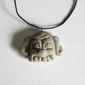 Handmade stone-textured ceramic pendant featuring a carved Taíno ancestral face petroglyph with a wide smile on a black cord necklace, inspired by indigenous Caribbean rock art.